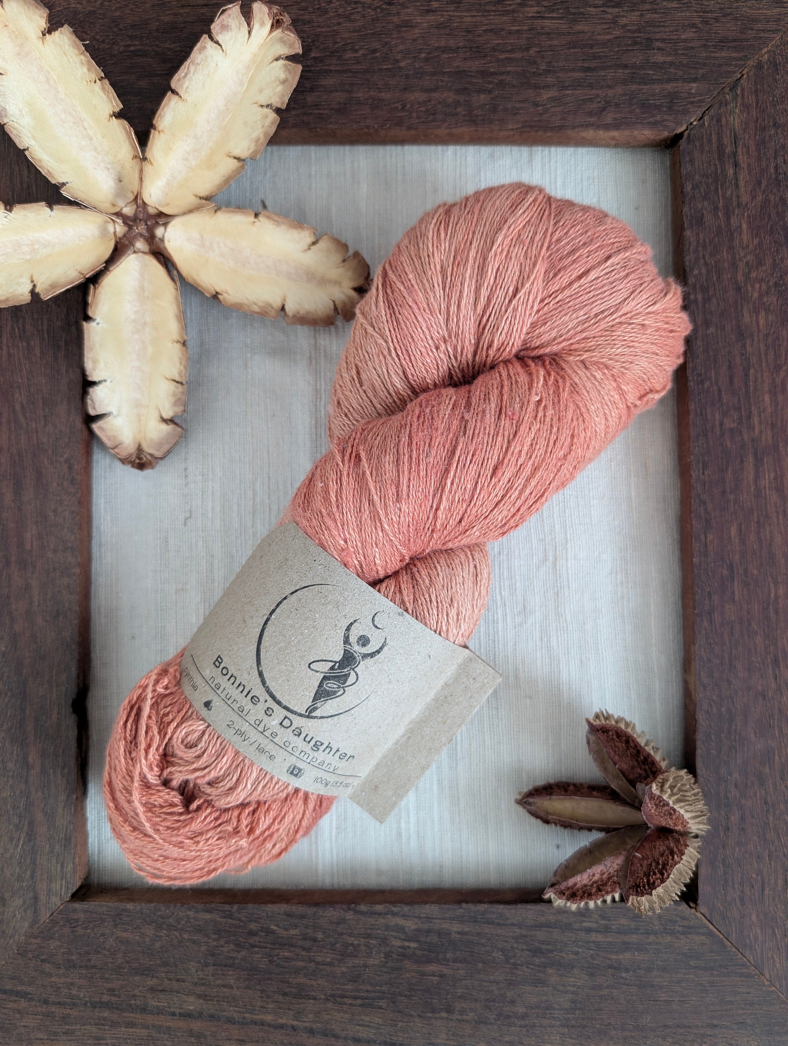 A naturally dyed skein of lace weight Eri silk yarn in tones of coral, salmon, and apricot. The skein is sitting on a piece of undyed Eri silk fabric, surrounded by a rustic wooden frame accented with seed pods of the Australian teak tree.