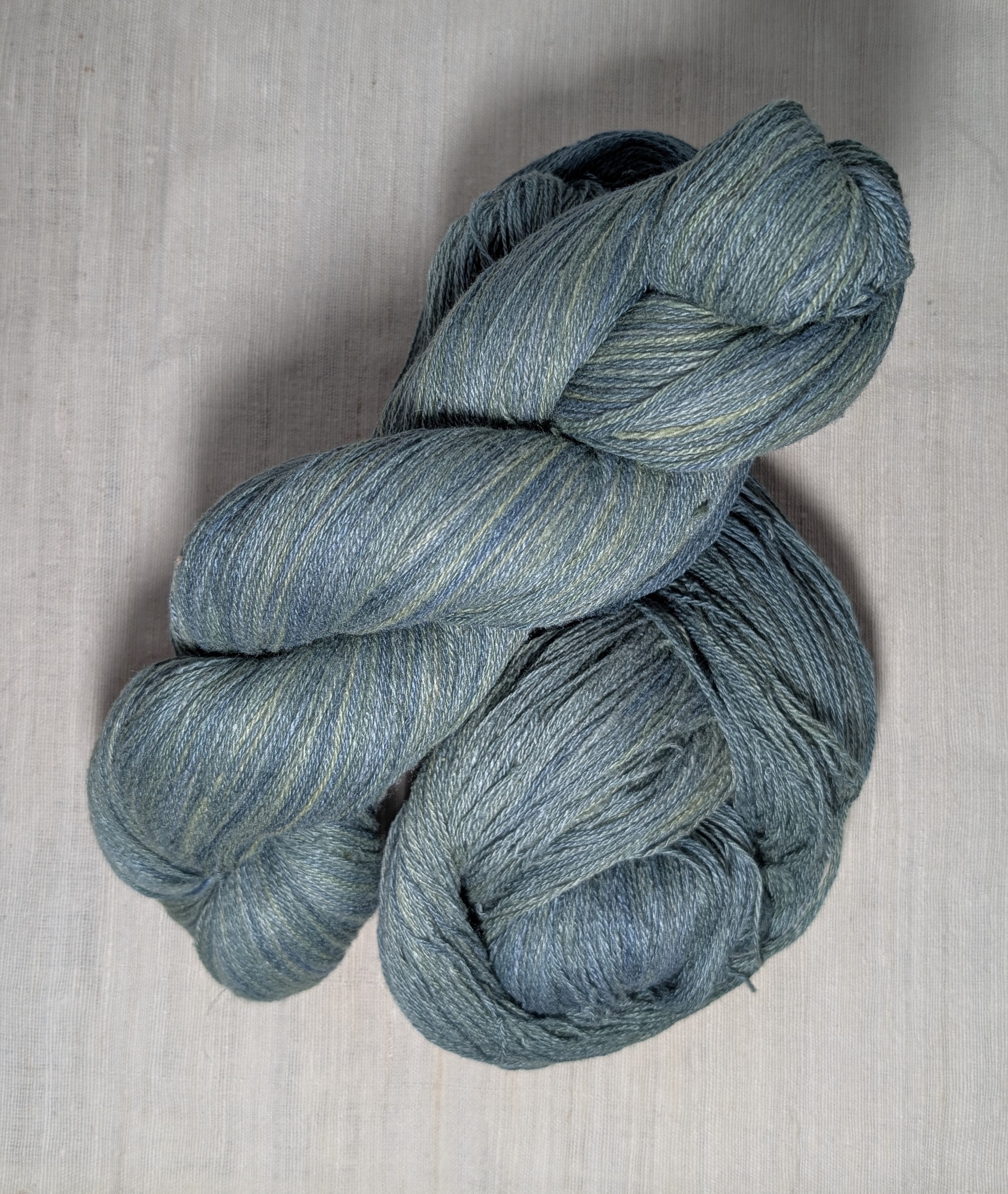 Two loosely twisted skeins of naturally dyed lace weight Eri silk yarn in an undulating teal colourway. The skeins sit on a piece of undyed hand-loomed Eri silk fabric.