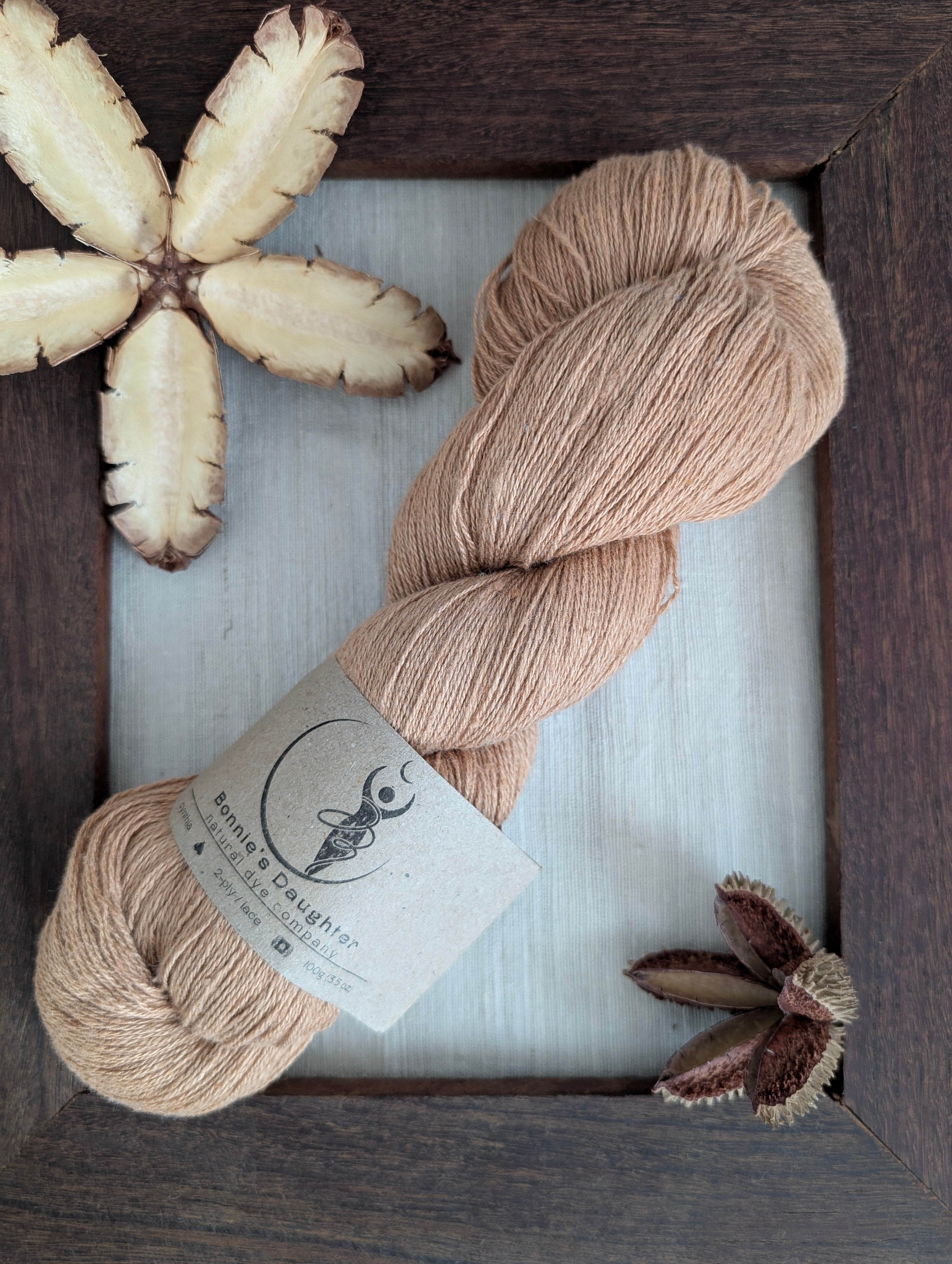 Warm golden amber coloured skein of naturally dyed Eri silk yarn with a label surrounded by a rustic wooden frame accented with seed pods of the Australian teak tree.