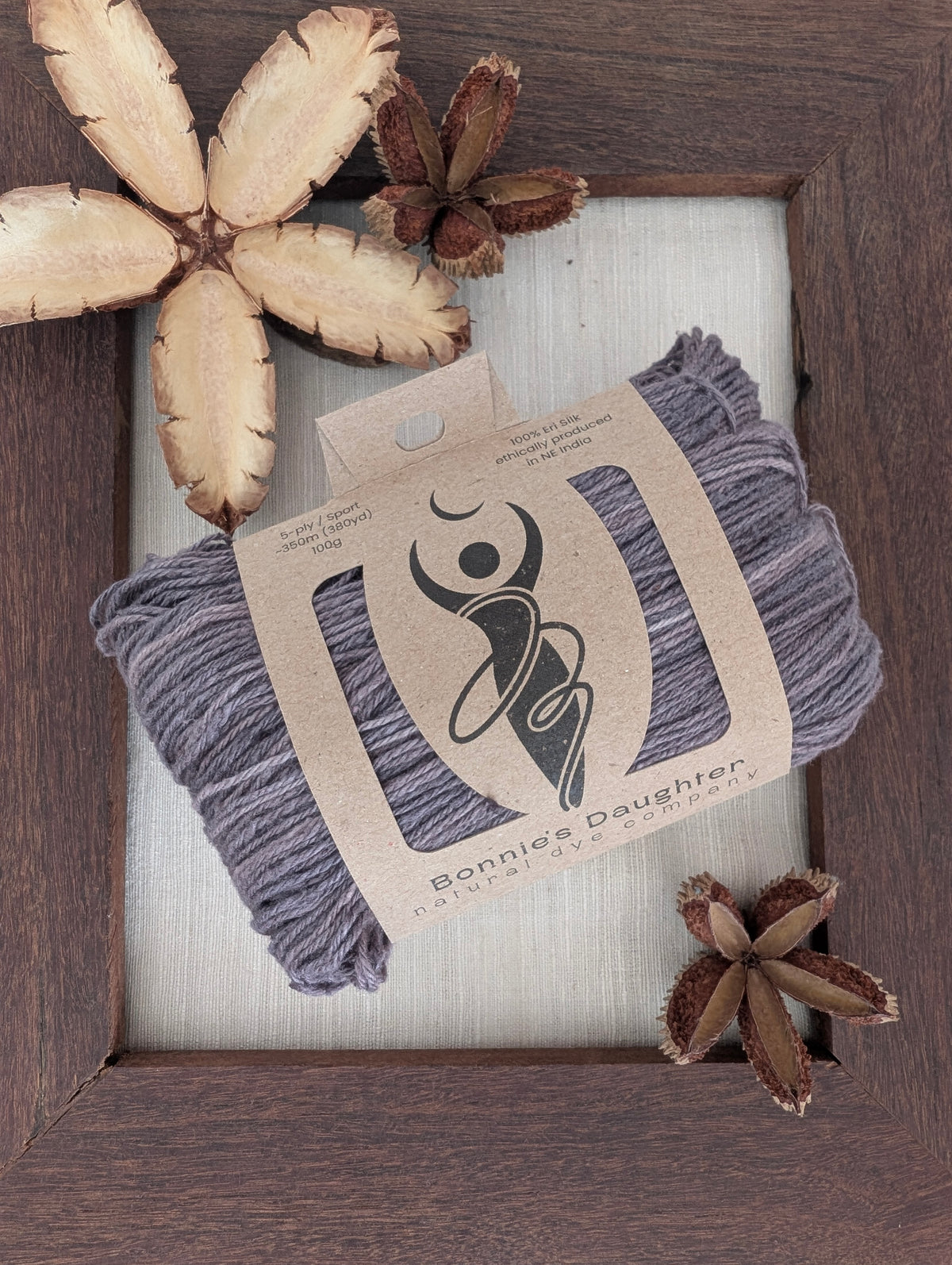 Smokey Amethyst 5-ply/Sport Eri silk yarn in smoky purple-grey tones, wrapped in a kraft label, on a piece of undyed Eri silk fabric in a rustic hand-made dark wooden frame accented with seed pods from the Australian teak tree