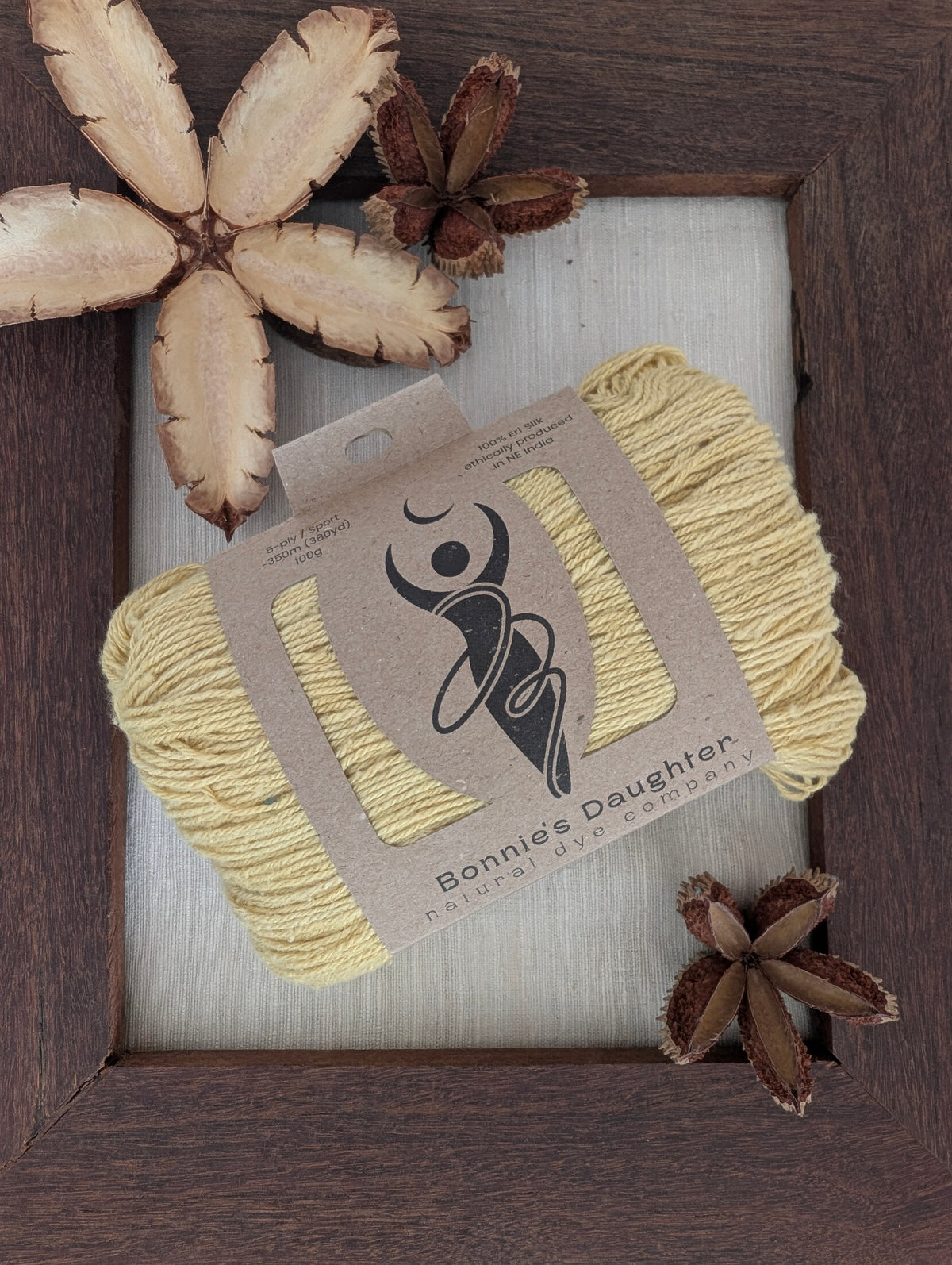 Solidago 5-ply/Sport Eri silk yarn in soft golden-yellow tones, wrapped in a kraft label, on a piece of undyed Eri silk fabric in a rustic hand-made dark wooden frame accented with seed pods from the Australian teak tree