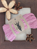 Luminous Orchid 5-ply/Sport Eri silk yarn in bright orchid-pink tones, wrapped in a kraft label, on a piece of undyed Eri silk fabric in a rustic hand-made dark wooden frame accented with seed pods from the Australian teak tree
