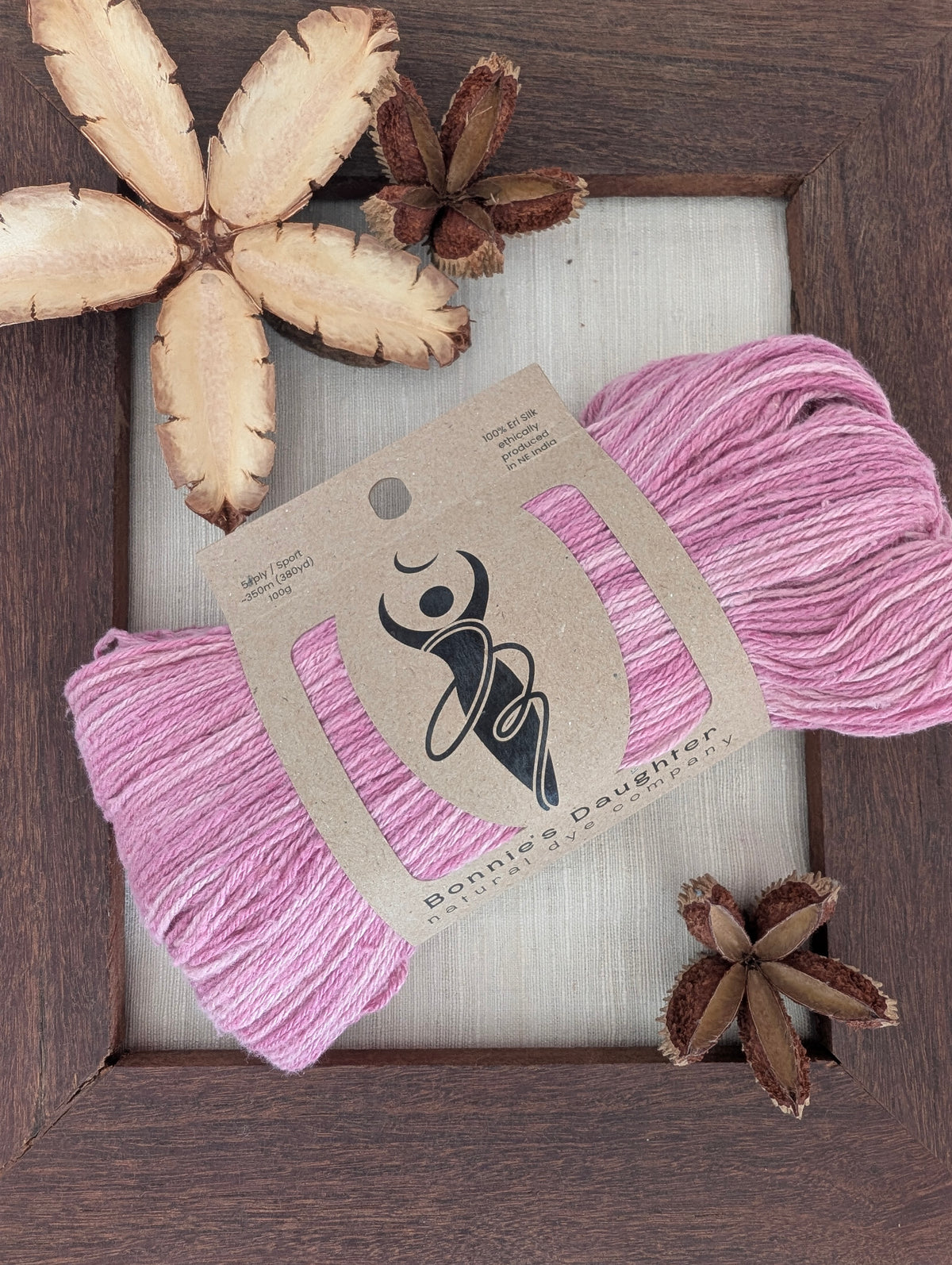 Luminous Orchid 5-ply/Sport Eri silk yarn in bright orchid-pink tones, wrapped in a kraft label, on a piece of undyed Eri silk fabric in a rustic hand-made dark wooden frame accented with seed pods from the Australian teak tree