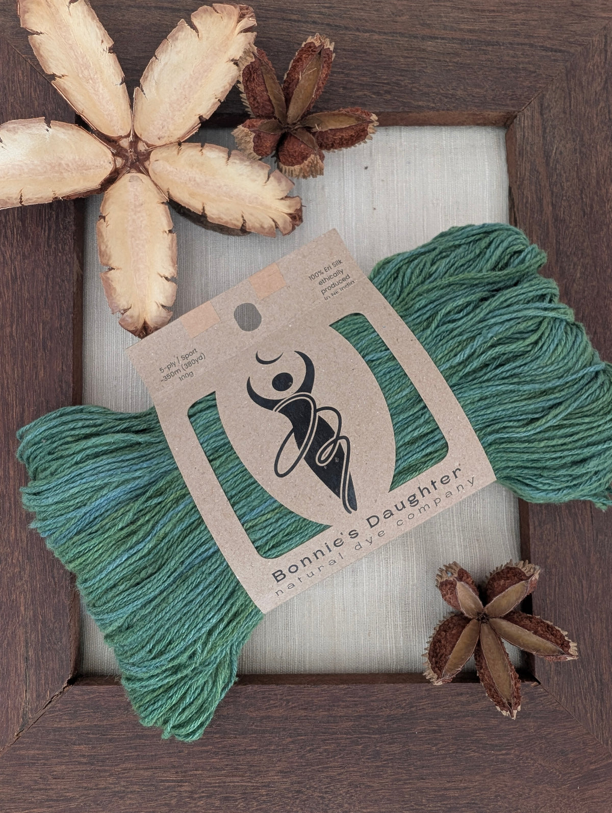 A skein of Bonnie's Daughter natural dye company teal green yarn with its kraft paper label, styled in a flat-lay on a white mat inside a dark wood frame with dried botanicals.