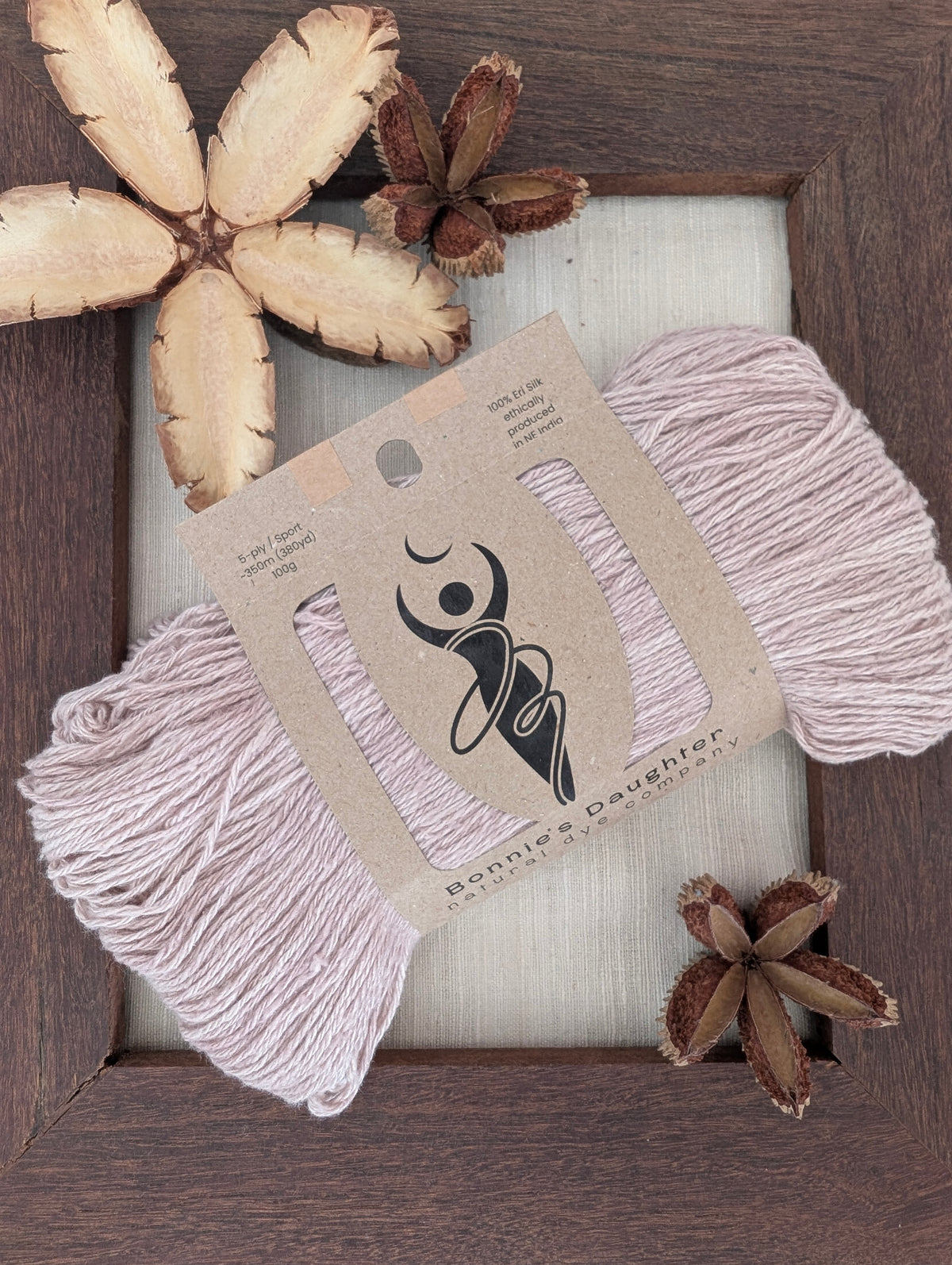 5-ply/Sport Eri silk yarn in pale lavender-grey, wrapped in a kraft label, on a piece of undyed Eri silk fabric in a rustic hand-made dark wooden frame accented with seed pods from the Australian teak tree⠋