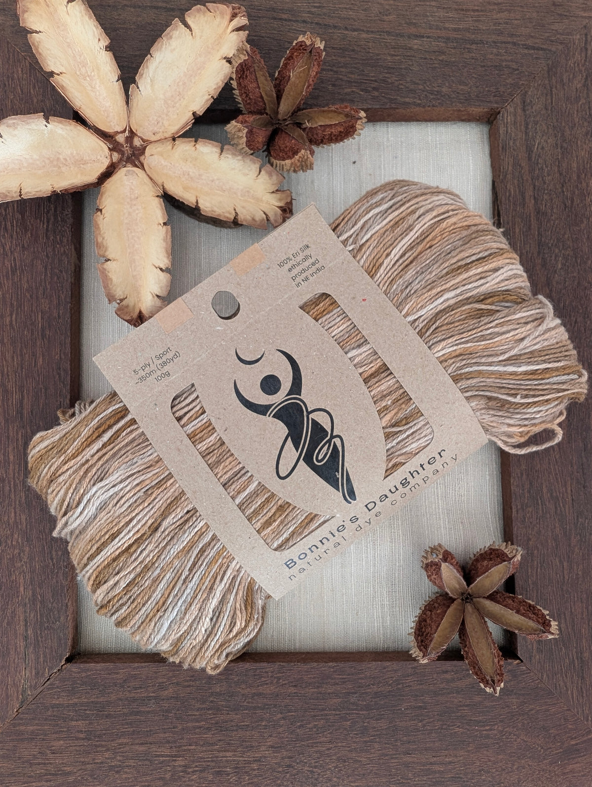 Imperial Topaz 5-ply/Sport Eri silk yarn in variegated brown, cream and grey, wrapped in a kraft label, on a piece of undyed Eri silk fabric in a rustic hand-made dark wooden frame accented with seed pods from the Australian teak tree