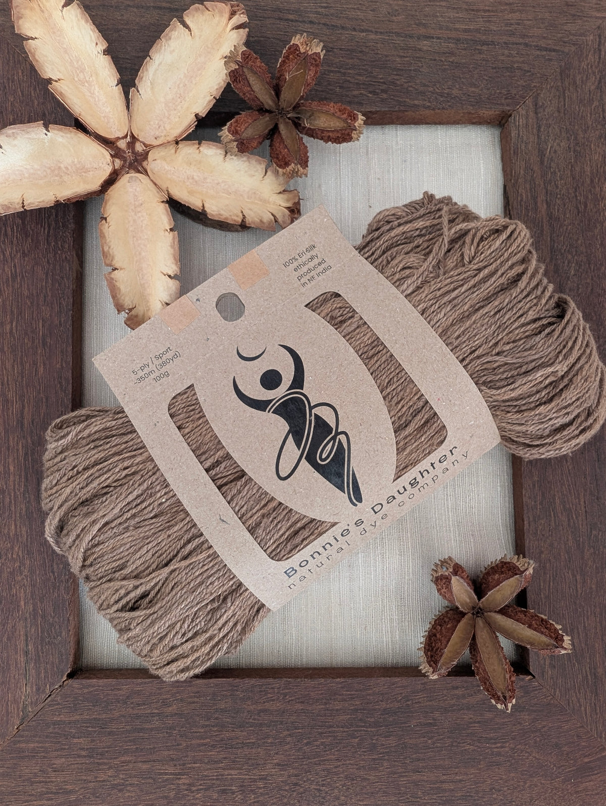Cocoa 5-ply/Sport Eri silk yarn in rich chocolate-brown tones, wrapped in a kraft label, on a piece of undyed Eri silk fabric in a rustic hand-made dark wooden frame accented with seed pods from the Australian teak tree.