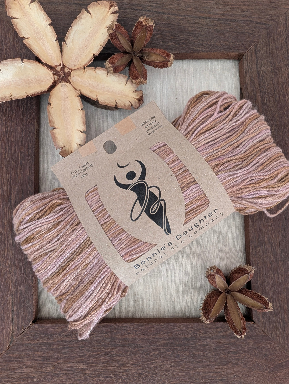 Laguna Agate 5-ply/Sport Eri silk yarn in variegated blush pink, dusty mauve and caramel-brown tones, wrapped in a kraft label, on a piece of undyed Eri silk fabric in a rustic hand-made dark wooden frame accented with seed pods from the Australian teak tree⠋