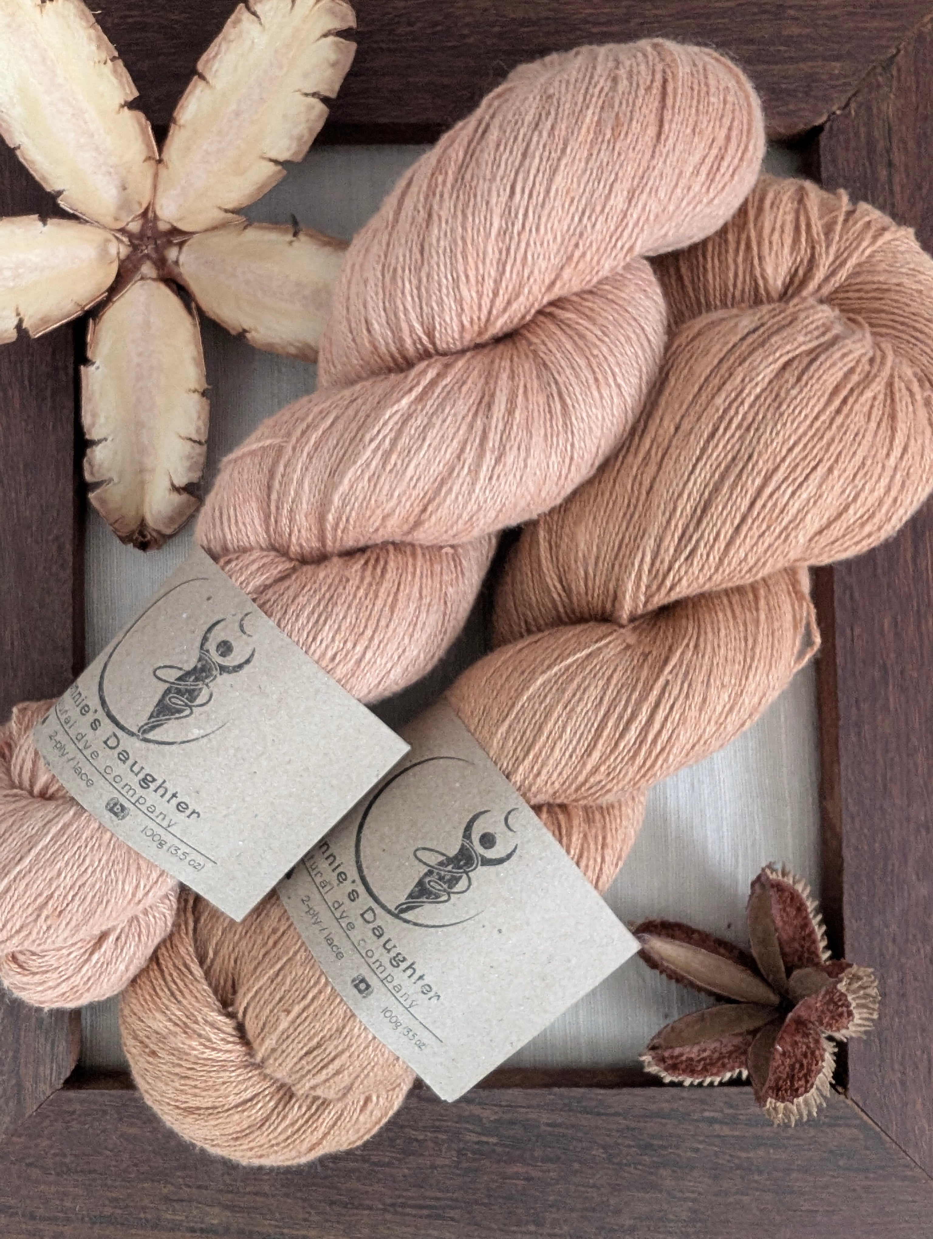 Two skeins of Eri silk yarn naturally dyed with Loquat leaf, showing the colour difference that can be achieved with this dye. The top skein is pinker in hue while the bottom one is a rich amber. The skeins sit atop a piece of hand-loomed Eri silk fabric and are surrounded by a rustic wooden frame, accented with seed pods of the Australian teak tree.